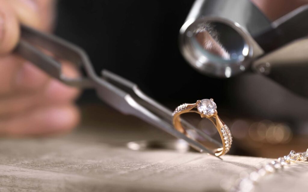 Goldring mit Diamanten der mit einer Pinzette gehalten und von einer Lupe betarchtet wird