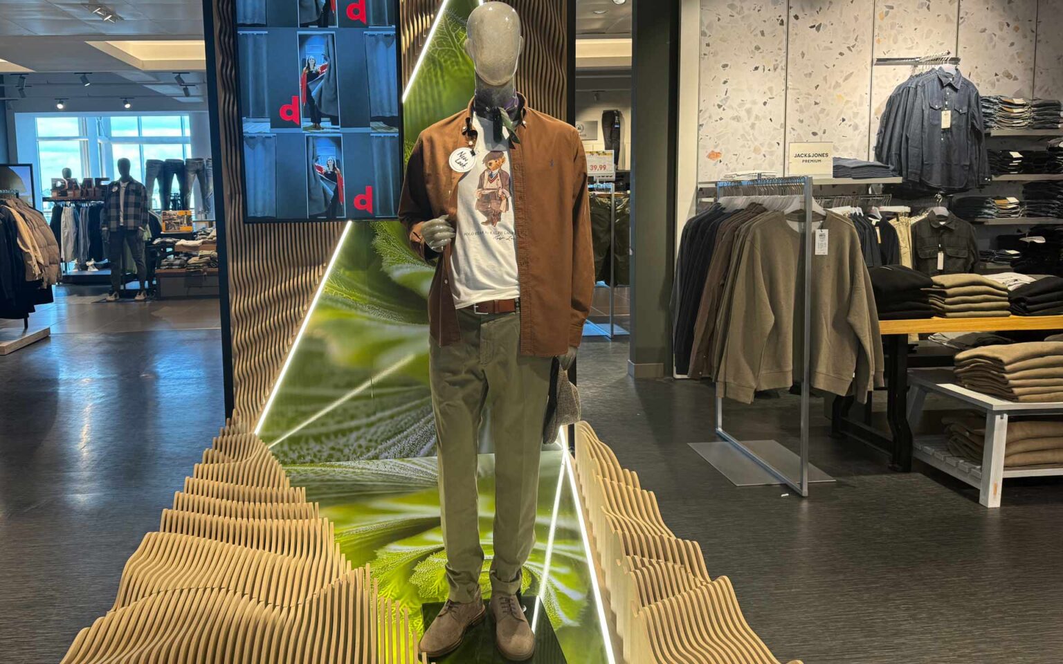 Puppe steht in der ModeWelt Herren und trägt eine Hose in der Farbe Olivgrün, Schuhe in taupe, ein Shirt in weiß, ein Hemd in braun und Accessoires in Grau-, Grün- und Brauntönen