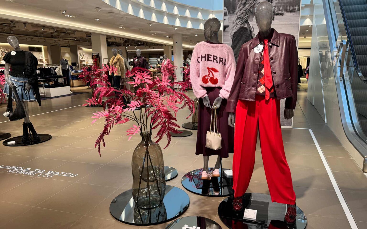 Highlightfläche an der Rolltreppe mit zwei Puppen im Vordergrund, welche herbstlich winterliche Outfits in den Farben Kirschrot, Weinrot und Rosa tragen.