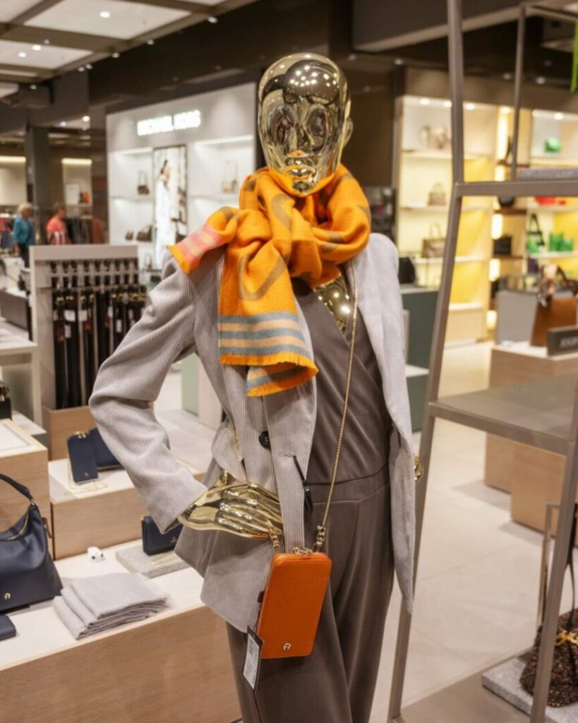 Damenfigur im Einteiler mit grauem Blazer, orangenem Schal und orangener Handytasche